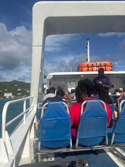 The image displays a forensic view from a passenger vessel in transit on the waters adjacent to Road Town, British Virgin Islands. The perspective is from the rear passenger seating area, looking forward towards the bow. Approximately 15 to 20 individuals, primarily adults, are visible as passengers; most are seated in blue canvas sling-style seats. The back of a passenger in a red shirt occupies the right foreground. A male individual, standing in the mid-cabin, wears a dark blue collared shirt with a logo and a dark baseball cap, holding a small white object in his hands. On the upper deck, aft of the main cabin, a white mast with navigation lights is present, along with two stacked orange cylindrical life rafts secured by netting. White structural components of the vessel, including railings, frame the scene. Signage on the cabin includes "NO SMOKING" and "RESTRICTED AREA." To the port side of the vessel, the clear turquoise water meets a verdant, hilly coastline featuring several scattered white buildings and a larger multi-story resort complex. The sky above is blue with scattered white cumulus clouds, indicating daylight conditions.