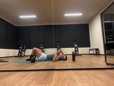The scene depicts an individual resting on an exercise mat inside a gym or fitness studio in Komchén, Mexico. The room features a light brown wood laminate floor, a large mirror wall spanning the length of the room, a dark gray or black accent wall, and a white wall. Overhead, a single rectangular fluorescent light fixture illuminates the space.
One person, appearing to be an adult, is lying supine on a teal exercise mat positioned centrally on the floor. They are wearing a gray short-sleeved shirt, light blue shorts, and black socks, with a purple resistance band positioned around their upper thighs. Their knees are bent, and feet are flat on the mat. The individual is holding a smartphone in their left hand, which is raised, while their right arm is bent with the hand near their head.
Adjacent to the person, on the mat, is a black kettlebell. A dark red water bottle rests on the floor to the individual's right. To the far left, off the mat, a pair of brown slip-on shoes are visible. Reflected in the mirror are three black stationary exercise bikes, positioned against the dark wall, indicating the presence of additional fitness equipment. A small black stool is visible against the white wall in the background to the right.