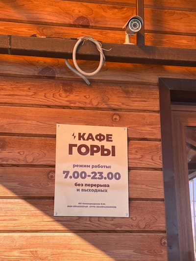 The image shows a close-up of a wooden exterior wall with a sign in Russian for "Cafe Gory" (meaning "Cafe Mountains"). The sign states the cafe's opening hours as 7:00 - 23:00 and indicates that it is open every day without breaks. The sign also includes the business registration information. A security camera is mounted on a post to the left of the sign with some exposed wiring. The location is Gory, Russia.