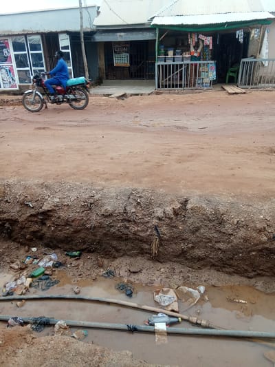 In Kagini, Nigeria, a motorcycle rider wearing a blue outfit navigates a dirt road beside a row of small shops. The road is unpaved and appears dry in some areas, but a trench filled with muddy water and debris is visible in the foreground. The trench contains discarded plastic bottles, bags, and other refuse, with pipes partially submerged in the murky water. The shops, characterized by corrugated metal roofs and open fronts displaying goods, line the side of the road. One shop features a sign with the word "LION" and images of products. Another shop appears to be a beauty studio with posters of faces on its exterior. The overall scene depicts a somewhat disorganized urban environment with evidence of ongoing infrastructure work or neglect in the form of the open trench and its contents.