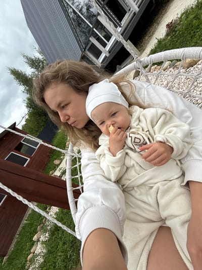 A young woman with blonde hair is sitting in a hammock, holding a baby. The baby is wearing a white beanie and a white hooded outfit, and is eating something. The woman is looking down at the baby. In the background, there is a wooden building and some greenery. The sky is cloudy. The location is Ryzhkovo, Russia.
