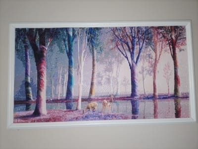 The image shows a framed diamond painting hanging on a plain, light-colored wall. The painting depicts a serene forest scene with a body of water. Tall, slender trees with colorful foliage dominate the composition. The trees are rendered in a mosaic-like style, with individual diamond drills creating a shimmering effect. In the foreground, two deer are grazing near the water's edge. The water reflects the trees and the soft, diffused light of what appears to be dawn or dusk. The overall color palette is a blend of purples, blues, pinks, and whites, creating a dreamy and tranquil atmosphere. There is no discernible location in the city of Kalisz, Poland, depicted in this artwork.
