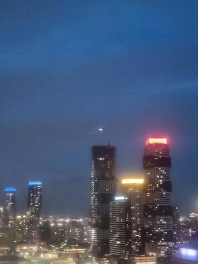The image shows a nighttime view of the Mississauga city skyline in Canada. Several skyscrapers are visible, with some under construction. One prominent skyscraper in the foreground is topped with bright red and orange lights. Another building nearby has a horizontal band of yellow light. To the left, two other buildings are illuminated with blue lights. The rest of the cityscape is dotted with numerous lights from buildings and streetlights, suggesting a densely populated urban area. The sky is a deep blue, indicating twilight or early night. There are no people or visible objects other than the buildings and lights in this scene.