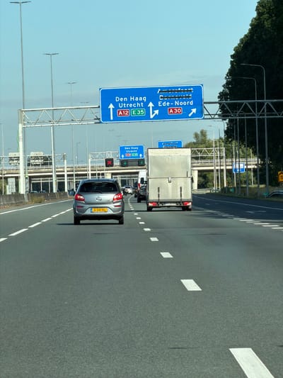 The image shows a highway scene with multiple lanes of traffic. A gray car with a license plate starting with "G-341-KL" is in the foreground, followed by a white truck. Several other vehicles are visible further down the road. Overhead signs indicate directions to "Den Haag Utrecht A12 E35" and "Amersfoort Corneld Ede-Noord A30". There are streetlights and trees lining the highway, and a blue sky above. The scene appears to be a typical highway environment. The sign indicating "Ede-Noord" places the location of the image in or near Ede, Netherlands.