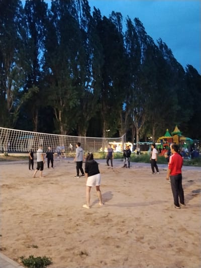 This media file shows a group of people playing volleyball on a sandy court. Several individuals are visible on the court, engaged in the game. One person in the foreground is wearing white shorts and a black top, with their back to the camera. To the left of the net, several other players are seen in various stages of the game, some with their backs turned, others facing the net. A person in a red shirt and dark pants stands on the right side of the court, facing away from the camera.

In the background, tall trees with dark foliage line the area, suggesting a park or recreational space. A volleyball net spans the width of the court. Further in the background, a brightly colored playground structure is visible, hinting at the location being a public area with facilities for both sports and children's activities.

The sky is a deep blue, indicating that the time of day is either early morning or late evening, likely dusk given the artificial lighting that appears to be present in some areas. The overall atmosphere seems relaxed and recreational. There is no visible text in the image. The location context provided is Bosteri, Kyrgyzstan.