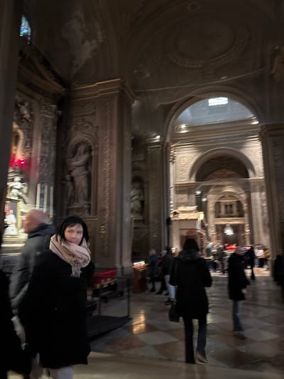 This image depicts the ornate interior of the Cattedrale di San Giorgio Martire (Ferrara Cathedral) in Ferrara, Italy. The scene shows a dimly lit, expansive religious space characterized by grand architecture.
In the foreground, a woman with dark hair, wearing a dark coat and a light-colored plaid scarf, is positioned slightly to the left of the center, looking directly at the viewer with a neutral expression. To her left, partially obscured, is a bald man wearing a dark jacket, facing away from the camera. A dark metal barrier or railing is visible in the lower left.
The midground and background reveal the cathedral's intricate details and other visitors. On the right midground, a woman with dark hair, wearing a dark coat, is seen from behind, walking away from the camera and carrying a dark handbag. Nearby, a stand holds multiple lit votive candles, with several individuals gathered around it. Further into the background, groups of people are dispersed throughout the large space, generally appearing to observe or move through the area.
Architecturally, the interior features tall, ornate pilasters lining the walls, each adorned with sculpted niches housing statues of robed figures. High arches lead into deeper sections of the cathedral, showcasing more elaborate decorations and recessed areas. The vaulted ceiling is visible, featuring decorative medallions and intricate plasterwork. On the far left, a prominent red glow emanates from what appears to be an altar or shrine, near which a figure in white vestments is discernible. The floor is covered with patterned tiles, likely marble or stone. The overall environment suggests a quiet, contemplative atmosphere typical of a historic cathedral interior.
