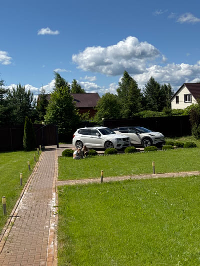 In the village of Borisovo DNP, Russia, on a sunny day, a red brick path leads through a well-maintained green lawn. Two white cars are parked on a paved driveway. A white BMW SUV is parked closer to the foreground, and a white electric car with a black roof is parked next to it. Two individuals, possibly a child and an adult, are sitting on the grass in front of the BMW. The background features several houses with dark roofs and mature trees, all under a bright blue sky with scattered white clouds. The scene suggests a peaceful suburban or rural setting.