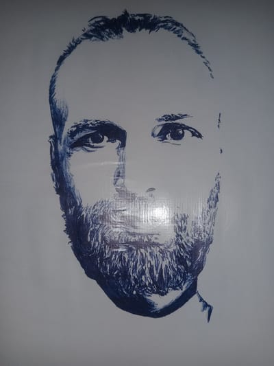 The image is a blue ink sketch of a man's head and shoulders against a white background. The man has a bald head with some hair on the sides, a beard, and is looking forward. The drawing is a sketch, not a photograph, and therefore does not depict any specific location, including Gulbag, Uzbekistan. The lighting in the image suggests it is an indoor setting, possibly a room with overhead lighting that casts subtle reflections on the surface of the drawing. The texture of the paper or surface on which the drawing is made is visible as a grid pattern.