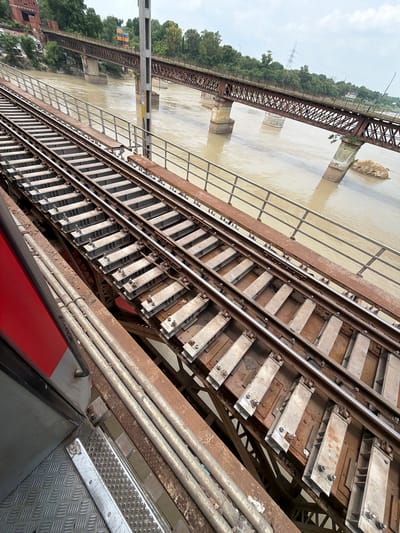 The image shows a train track bridge over a murky river. The bridge is made of rusted metal beams and concrete supports. There are two sets of train tracks on the bridge. The train tracks are parallel and run diagonally across the image. The river is wide and brown, with some debris floating in it. The sky is overcast. The location is Shuklaganj, India. In the foreground, part of a red train car is visible.
