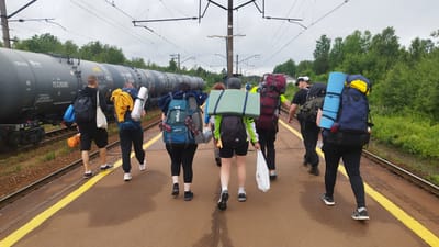 A group of eight individuals, each carrying large backpacks, are walking away from the camera on a train platform in Orekhovo, Russia. The platform is paved with asphalt and has a yellow line marking the edge. To the left of the platform are train tracks and a long, dark-colored train composed of multiple carriages. The train carriages appear to be tankers, with visible markings and numbers, including "75328385". Overhead, power lines for the train are visible. The background consists of dense green trees and foliage, with a cloudy sky overhead. The individuals are dressed in casual clothing suitable for travel or outdoor activities. Some are wearing hats and headphones. The backpacks are of various colors and sizes, some with sleeping mats or other camping gear attached. Some individuals are also carrying bags or bottles. The scene suggests a departure or arrival of a group on a train journey.