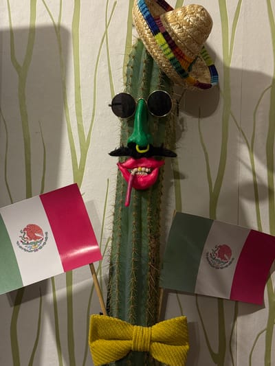The image displays a flat-lay arrangement of objects with a Mexican theme. A straw sombrero with a multicolored band lies to the left. A green cactus with visible spines stretches across the center of the image. On the cactus are sunglasses, a red and green painted figure, and two Mexican flags on sticks. A yellow woven bow tie is on the right. The objects are placed on a patterned surface with horizontal lines. The location is not identifiable as Ketoshi, Russia, and no people, actions, or interactions are depicted.