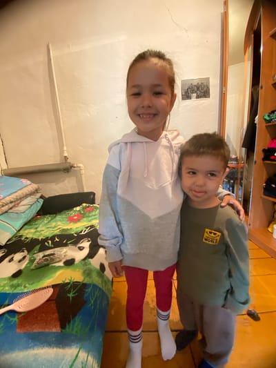 Two children, a girl and a boy, stand together in what appears to be a bedroom. The girl, taller, is on the left, wearing a pink and gray hoodie and red leggings with white socks. Her hair is pulled back in a ponytail, and she is smiling. The boy, on the right, wears a green long-sleeved shirt with a yellow graphic on it and gray pants. He is sticking his tongue out slightly. They are positioned in front of a bed with colorful bedding featuring cartoon characters, and a pink hairbrush is visible on the bed. Behind them, the wall is a pale color with some visible cracks, and a pipe runs horizontally across it. A framed black and white photograph of several people is hanging on the wall above the pipe. To the right, a wooden closet or shelf unit is visible, with clothing items stacked on shelves. The flooring is a warm-toned, possibly wooden, surface. The scene is dimly lit, suggesting an indoor setting during the day. The location is identified as Maloarslangulovo, Russia.
