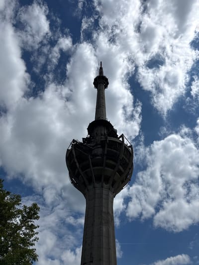 An upward shot of the Avala Tower in Sremska Kamenica, Serbia. The tower's concrete support structure is visible, ascending toward its distinctive latticed, rounded section. The camera angle emphasizes the tower's height. The background is a sky with cumulus clouds and patches of blue sky. Trees with green leaves are visible in the lower right corner.