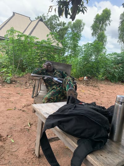 In the city of Sankara, Nigeria, an outdoor scene depicts a male individual in a camouflage uniform resting. He is seated on a wooden chair, with his right hand supporting his head and eyes closed. An assault rifle, consistent with an AK variant, lies across his lap. His left foot, wearing a combat boot, is propped on a lower wooden bench in the foreground. A black backpack and a silver thermos are also present on this bench. The uniform bears a partially visible name patch reading "GOODLUCK". The ground is reddish-brown dirt. In the background, dense green vegetation, including trees and shrubs, partially conceals a building with a light-colored, multi-gabled roof. Two chickens, one brown and one white, are visible among the vegetation in the mid-ground. The sky is overcast.
