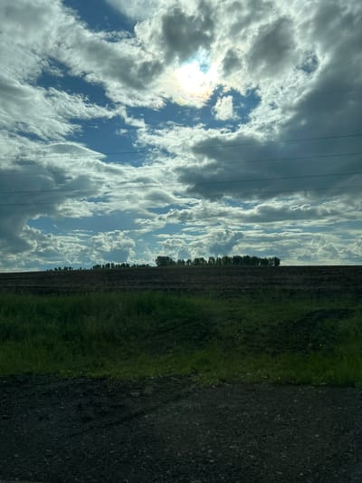 This image, captured in Sukhtelinskiy, Russia, depicts a broad rural or agricultural landscape under a partly cloudy sky. The upper two-thirds of the frame are dominated by a sky featuring a mixture of bright white cumulus clouds and darker, more stratified cloud formations, interspersed with patches of clear blue sky. The sun is partially obscured by clouds in the upper-left quadrant, producing a bright glare with visible spectral hues. Three parallel horizontal utility lines (power lines) traverse the sky across the middle section of the image.

A distinct horizon line separates the sky from the land. Below the horizon, a dense, dark band of trees or shrubbery stretches across the width of the image. In the mid-ground, a wide expanse of dark, tilled earth occupies a significant portion of the lower frame, appearing as a fallow or recently plowed field, devoid of visible crops. The immediate foreground consists of a band of dark green, somewhat overgrown grass, which transitions into a darker, granular surface, likely a gravel road or dirt track, at the very bottom of the image.

No people, animals, or active human interactions are visible within the frame. The lighting indicates daytime, with the sun's position behind clouds creating diffused light and localized bright spots where it penetrates the cloud cover.