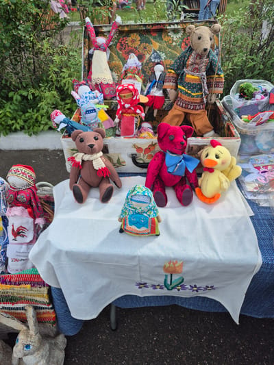 The image depicts a street market scene in Kansk, Russia. A collection of handmade fabric dolls and stuffed animals are displayed on a white tablecloth draped over a table. The dolls exhibit traditional Russian folk attire, with some featuring elaborate headscarves and embroidered patterns. Among the collection are several teddy bear figures, a yellow chick plush toy, and other assorted fabric creations. In the background, a vintage suitcase serves as a backdrop for some of the dolls, adorned with a colorful floral pattern. Beyond the immediate display, foliage and hints of people in casual attire suggest an outdoor setting. The ground appears to be asphalt, characteristic of a public street or square.