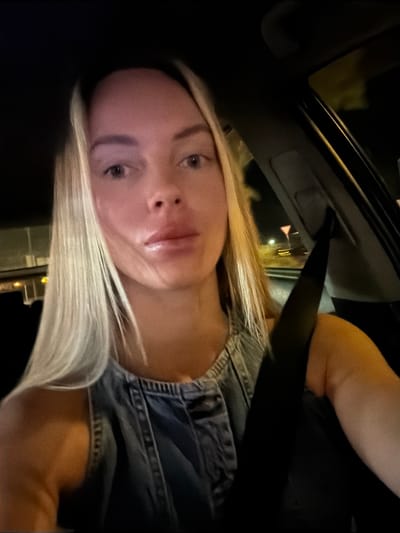 A blonde woman is pictured from the chest up, seated in the driver's seat of a car at night, likely in La Cala, Spain. She is wearing a denim-like top and her seatbelt is fastened across her chest. The background is dark, with some blurred lights suggesting a roadway or city lights visible through the car window. Her face is illuminated by the interior or external light, and she appears to be looking towards the camera. The overall scene conveys a sense of movement and nighttime ambiance.