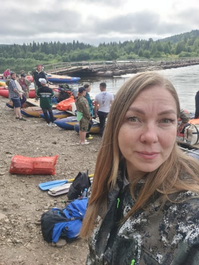 The image depicts a group of people on a rocky riverbank, with several inflatable rafts and a makeshift wooden bridge in the background. The scene appears to be outdoors in a natural setting, possibly a park or recreational area in Вишерогорск, Russia, given the user's prompt. The sky is overcast, suggesting it may be a cloudy day.
In the foreground, a woman is taking a selfie. She has long blonde hair and is wearing a camouflage jacket. Behind her, several other individuals are visible, some standing and others near the rafts. One person in a green jacket with "ЭКЛОС" written on it stands next to a raft.
The riverbank is covered in pebbles and small rocks. Several large, red, tarp-wrapped objects are present, possibly containing supplies or equipment. Various backpacks and outdoor gear, including paddles, are scattered around. In the middle ground, a long, wooden pontoon bridge crosses the river, connecting the two banks, which are lined with dense green trees. The overall atmosphere suggests a group preparing for or returning from an outdoor activity, likely involving watercraft.