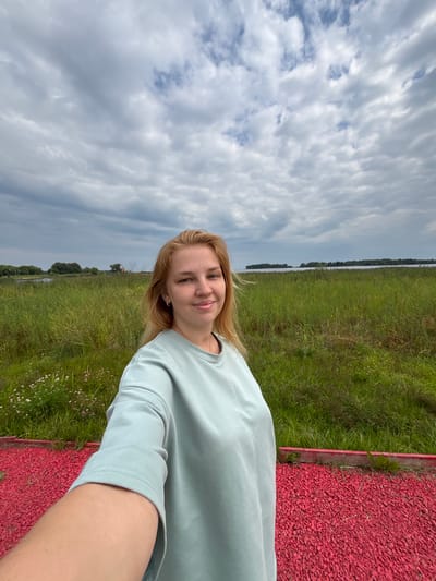 A young woman with blonde hair and light skin is taking a selfie in an outdoor setting. She is smiling and looking at the camera. She is wearing a light blue long-sleeved shirt. In the background, there is a field of tall green grass and a body of water, likely a lake or river, with a treeline in the distance. The sky is cloudy. In the foreground, there is a red gravel path. The image appears to have been taken in the village of Breytovo, Russia, given the typical landscape and architecture often found in such rural Russian locations.