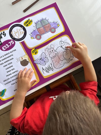 A child is coloring on a placemat at a table. The placemat has a menu on the left side and coloring activities on the right, including a connect-the-dots car and a drawing of snails and a butterfly. The child is wearing a red shirt and has their arm extended, holding a red crayon to color one of the snails. The background shows a white tabletop and wooden chair legs. The scene is likely in a restaurant.