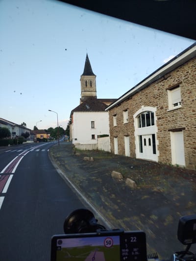 The image shows a view from inside a vehicle looking out through the windshield. The scene depicts a road in a village, likely in Firbeix, France, given the architectural style and the presence of a church spire.

In the foreground, a GPS device is mounted on the windshield, displaying a navigation map and speed information, including "50" inside a red circle, likely indicating a speed limit. The road ahead has white and red lane markings and a pedestrian crossing.

To the right of the road, a stone building with a white-framed arched doorway and double white doors is visible. Above this, a white-framed window with closed shutters is present. Further along the street, another stone building can be seen with white-framed windows and doors.

On the left side of the road, residential houses with light-colored facades are lined up, some with balconies and manicured greenery.

In the background, a prominent church with a tall, dark spire dominates the skyline. The sky is clear and light blue, with a few scattered clouds, suggesting it is late afternoon or early evening. The overall environment appears to be a quiet, semi-rural setting within a village. There are no people visible in the image.