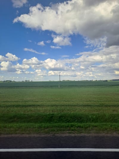 The image depicts a wide, flat agricultural landscape under a partly cloudy blue sky. In the foreground, a dark asphalt road with a white line runs horizontally across the bottom of the frame. Behind the road is a strip of green grass, followed by a vast field of green crops, likely wheat or barley, with visible rows. In the mid-ground, a thin, solitary utility pole stands in the field. The horizon is marked by a line of trees and vegetation. The sky above is filled with a mixture of white, fluffy cumulus clouds and darker, more substantial gray clouds, suggesting dynamic weather conditions. The scene appears to be captured from a moving vehicle, indicated by the slight blur in the foreground vegetation and road. The location is identified as Свіслацкі сельскі Савет, Belarus.