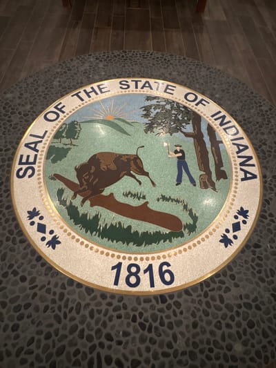 The image shows a circular seal embedded in a floor made of small, dark gray stones. The seal is the "Seal of the State of Indiana" and features a central design of a bison jumping over a log, with a frontiersman chopping down a tree in the background. A sunrise with rays is visible above rolling hills. The text "SEAL OF THE STATE OF INDIANA" encircles the upper portion of the seal, and the year "1816" is prominently displayed at the bottom. The seal is bordered by a thin gold ring and adorned with blue floral or star-like emblems on either side. The flooring around the seal is consistent with the surrounding dark stone. The image does not provide any specific information or visual cues that indicate its location within New Harmony, United States.