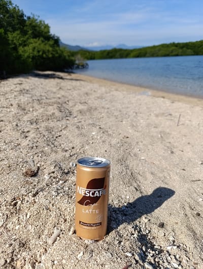 A can of Nescafe Latte is lying on a sandy beach in Kecamatan Banyuputih, Indonesia (latitude: -7.754, longitude: 114.367). The can is partially obscured by its own shadow. The beach is made of fine sand, and there are some small rocks and shells scattered around. In the distance, there is a body of water. The water is calm and appears to be a bay or lagoon. Beyond the water, there are low hills in the distance covered in vegetation. The sky is blue with a few white clouds. The scene is peaceful and tranquil.