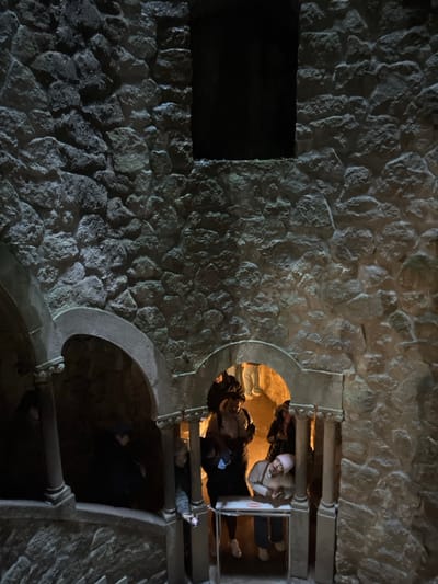 An unusual view of people in a stone structure, possibly a well or tower. The main wall is made of rough-hewn stones with a dark, recessed rectangular area on the left. To the right, a circular opening reveals people peering out, and a lower arched opening leads into deeper darkness. The overall lighting is dim, emphasizing the texture of the stone. The perspective gives the sense of looking up from below.