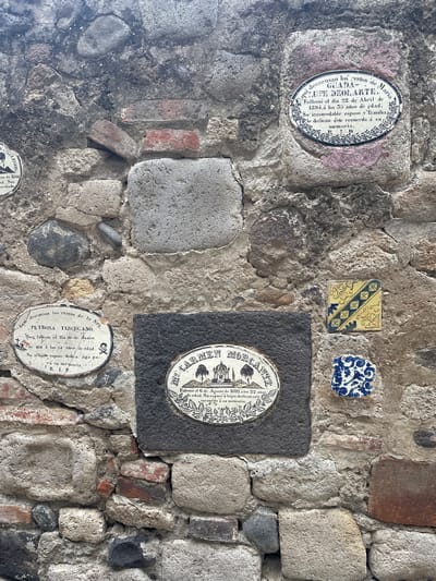 The image shows a section of a stone and brick wall with several commemorative plaques and tiles affixed to it. These appear to be part of a historic structure, possibly a cemetery or church in Huejotzingo, Mexico.

The central focus is a dark gray rectangular stone on which an oval plaque is mounted. This plaque features a decorative border surrounding an illustration of a domed building with trees and people. Below the illustration, text is inscribed in Spanish, indicating a memorial for someone named "Ma. del Carmen Mortante," who died on August 6, 1892, at the age of 22. Her husband and children dedicated this memorial.

To the left of the central plaque is another oval plaque, white with black lettering and a decorative border. This plaque memorializes "Petrona Tuscugano," who died on June 29, at the age of 34. Her afflicted husband dedicated this to her memory, with "R. I. P." inscribed at the bottom. A small skull and crossbones symbol is also present on this plaque.

Above the "Petrona Tuscugano" plaque, and slightly to the right, is a third oval plaque, white with a black border and decorative elements. This plaque memorializes "Guada-Lupe Deolart," who died on April 22, 1894, at the age of 35. Her inconsolable husband and family dedicated this to her memory.

Interspersed among the plaques are various tiles. Two small square tiles are visible in the upper right quadrant: one with a yellow background and a blue diamond pattern, and another with a blue and white floral design.

The wall itself is constructed from irregular-shaped stones, some of which are rounded river stones, and portions of red brick. The mortar between the stones is visible and shows signs of age and weathering. The overall impression is one of a well-maintained historical site.
