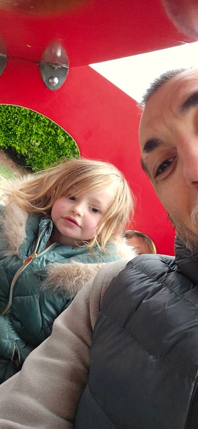 A selfie photograph taken from a low angle shows a man with a beard, and a young girl with blonde hair, both wearing winter jackets. The man's jacket is black and quilted, while the girl's jacket is teal with a fur-trimmed hood. They are positioned in front of a bright red structure with a circular cutout showing a patch of green vegetation. The structure is part of a playground installation, indicated by the presence of metal fixtures. The photograph was taken at Eaton Park in Norwich, United Kingdom.