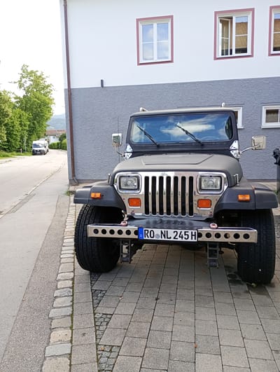 A dark gray Jeep Wrangler is parked on a cobblestone sidewalk in Brannenburg, Germany, in front of a two-story building. The building has a white upper story with red-trimmed windows and a gray textured lower story. The Jeep has a chrome grille, square headlights, and a chrome bumper with circular cutouts. Its license plate reads "RO NL 245 H". In the background, a road is visible with a white van and trees lining the side. The sky is cloudy.
