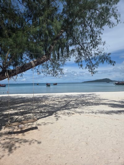 The image depicts a serene beach scene in Koh Toch Village, Cambodia. In the foreground, a simple wooden swing hangs from a large tree branch, casting dappled shadows on the white sand. The swing is unoccupied. Beyond the sandy shore, the calm, turquoise ocean stretches out to the horizon. Several small fishing boats are visible in the distance, dotting the water. The sky above is a bright blue, with scattered white clouds. The overall atmosphere is peaceful and tranquil, suggesting a perfect day for relaxation by the sea.