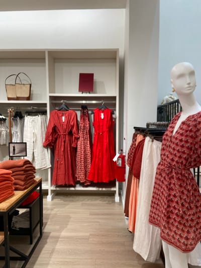 The image shows the interior of a clothing store in Greenwich, Connecticut. The location's coordinates are latitude: 41.029, longitude: -73.626. The store is brightly lit and has white walls and wooden floors. Several racks of clothing are displayed, with a variety of dresses and other items for sale. One mannequin is on display wearing a red, printed dress with a white background. The mannequin's face is painted white. The other dresses on the rack are all red. There is a large wicker bag in the top shelf of the left-hand rack, which is also white. The space is organized, and many garments are on display. The clothes are displayed in a modern and stylish way, with a focus on color and texture. The image suggests a high-end clothing store that caters to a discerning clientele.