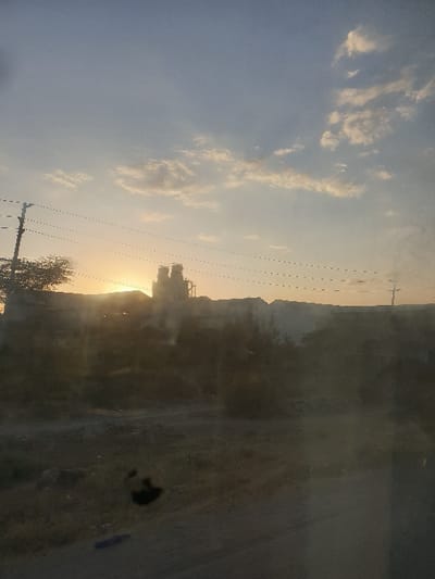 The sun is setting over a landscape featuring structures resembling a factory or industrial complex. The sky is filled with clouds, transitioning from a bright light near the horizon to a pale blue higher up. Power lines stretch across the scene, connecting the industrial area to the rest of the world. On the right side of the image, there's a blurred or obscured view of what seems to be a roadside or ground area.