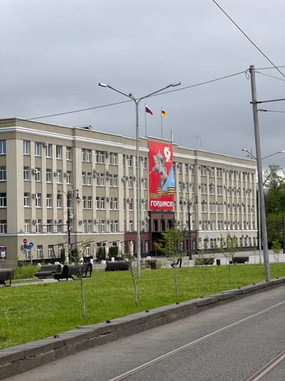 A vertical shot shows a tall building with many windows and air conditioning units, situated beside a patch of green grass and a street with tram tracks. A large red banner with Russian text and imagery of a statue hangs on the building. There are two flags atop a pole. The sky is overcast. The location is unknown.