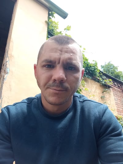 This is a selfie of a man with a shaved head and a short mustache, looking directly at the camera. He is wearing a dark blue sweatshirt. The background shows a wall with some peeling paint and ivy growing on it, suggesting an outdoor setting. Further in the background, there is a brick wall and some trees. The lighting suggests it is daytime, possibly overcast, as there are no strong shadows. The man's expression is neutral, with his eyebrows slightly furrowed. The image appears to be taken in Sloviansk, Ukraine.