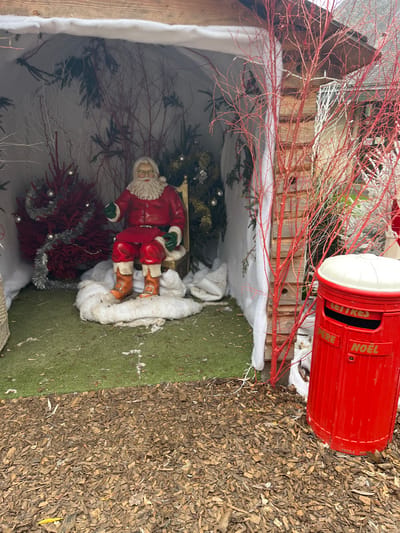 This image captures an outdoor Christmas display in the city of Bagnoles de l'Orne Normandie, France. The scene features a seated Santa Claus figurine, depicted with a long white beard, spectacles, a red suit, green gloves, and brown boots, positioned on a golden-colored chair. The Santa figure is surrounded by a small accumulation of artificial white snow, which rests on green artificial turf.
Behind Santa, the display's backdrop consists of white fabric draped to resemble snow, interspersed with numerous bare tree branches, some painted red and others white. Several artificial Christmas trees are visible; one on the left is red and adorned with silver tinsel and spherical ornaments, while others are green with similar embellishments.
The display is housed within an open wooden structure, identifiable by a visible wooden beam across the top and vertical wooden planks forming a side wall to the right. In the foreground, the ground is covered with dark brown wood chips. To the right, immediately outside the wooden structure, stands a bright red letterbox with a white domed lid. The letterbox has a horizontal mail slot and is inscribed with "LETTRES PÈRE NOËL" (Letters Father Christmas) in gold lettering. Additional red-painted branches are visible to the right, adjacent to the letterbox and the wooden structure.