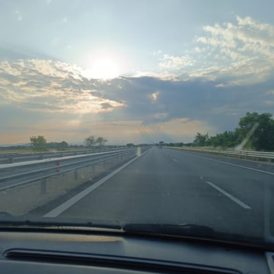 The scene depicts a multi-lane highway viewed from inside a moving vehicle, located in the vicinity of Saraia, Bulgaria. The sky occupies the upper two-thirds of the image, featuring a low, bright sun positioned in the upper-left quadrant, partially obscured by numerous clouds. Crepuscular rays (sunbeams) fan out prominently from behind the clouds, extending across the sky towards the right. The clouds vary in formation, from scattered cumulus-like structures to more continuous, darker strata.
The highway extends into the distant horizon, marked by clear white lane lines. Several light-colored vehicles are visible ahead, traveling in the same direction. Metal guardrails run parallel to the road on both the left and right sides. Beyond the guardrails, the landscape includes green trees, some appearing as clusters, and sections of lighter-colored vegetation or fields. Distant hills or mountains are faintly visible on the horizon line.
The bottom portion of the image shows the dark dashboard and the base of a windshield wiper, indicating the perspective is from the interior of the vehicle, likely the driver's or front passenger's seat. A subtle reflection or glare is visible on the windshield in the lower-right area. No individuals are directly visible in the image.