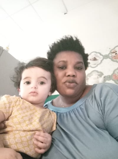 This image depicts an adult female and an infant in a close-up, indoor portrait, likely taken in Basra, Iraq. The adult female, appearing to be of African descent, has dark skin and short, dark, curly hair. She is wearing a light blue/teal short-sleeved top with a gathered neckline and a thin necklace, looking directly at the camera with slightly pursed lips. The infant, positioned to the adult's left and appearing to be of lighter complexion with dark, wavy hair, is wearing a yellow short-sleeved top with a subtle patterned print. The infant also looks directly at the camera with a neutral expression, its left hand resting on the adult's upper arm or shoulder.
The background reveals details of an interior setting. To the left of the subjects, a dark gray or metallic object, possibly a refrigerator or cabinet, is partially visible, with a yellow bottle resting on its surface. To the right, a wall is adorned with decorative wallpaper or tiles featuring repeating light green, cloud-shaped motifs, each containing an illustration of household items such as teacups and teapots. The ceiling above is white with visible grid lines. The scene is brightly lit, with some areas appearing overexposed. The interaction suggests a familial relationship.
