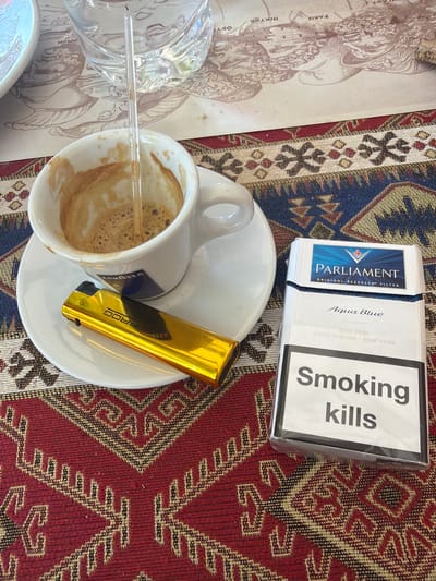A close-up, overhead shot shows a table setting with a partially consumed espresso in a white cup, a gold lighter, and a pack of Parliament Aqua Blue cigarettes. The items are placed on a patterned rug. In the background, a glass of water sits on a placemat with a historical illustration that appears to depict figures from Greek mythology and text labels including "PARIS", "HEKTOR", and "ODYS". The scene is likely set in a cafe or restaurant in the Çanakkale Merkez area of Turkey, given the visual cues and common cafe offerings.