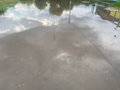 The image depicts a flooded area, likely after heavy rainfall, with murky water covering the ground. In the reflection on the water, a tall, lattice-like tower, possibly a radio or cell tower, is visible. A utility pole with wires is also reflected. On the right side of the reflection, a fence and some yellow structures, which could be buildings or sheds, are discernible. The surrounding environment appears to be a mix of vegetation and some form of urban or semi-urban infrastructure. Small white specks are scattered across the surface of the water. The overall scene suggests an outdoor location in a low-lying area that has become inundated with water. There is no visible indication of the specific location within Заволжский, Russia.