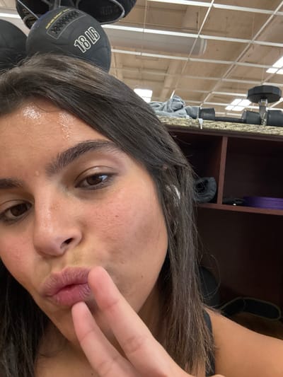The image captures a close-up selfie of a female individual within a gym setting in Plainsboro Township, United States. The individual has dark brown hair and displays visible perspiration on her forehead, indicative of physical exertion. Her lips are pursed, and her right hand is positioned near her mouth with the index and middle fingers extended in a "V" shape. In the background, two black medicine balls are partially visible, one marked "18 LB". To the right, a granite-patterned counter holds dumbbells and a grey towel. Below the counter, a dark brown shelving unit contains items, including purple mats and a black object. The ceiling features a grid of white acoustic tiles.