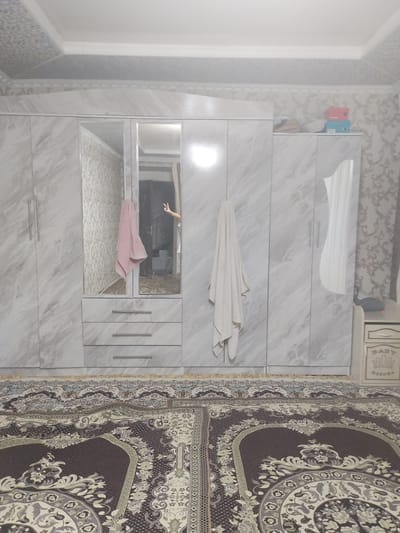 A bedroom scene in Kyrgyzstan, during daytime. The room is furnished with a large, modern wardrobe with a marble-effect finish. The wardrobe has multiple doors and drawers, and features a mirrored section. Two towels are visible, one pink and one white, hanging from the wardrobe. A person's hand is visible in the mirror, giving a peace sign. On top of the wardrobe, two colorful boxes are stacked. To the right of the wardrobe, a small nightstand with decorative carvings is present. The floor is covered with a patterned rug. The ceiling is decorated with patterned panels and molding. The overall impression is of a tidy and organized room, likely a personal living space. The style of the furniture and decor suggests a contemporary aesthetic within a socialist context, common in Kyrgyzstan. There is no visible text in the image.