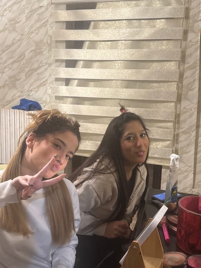 This image depicts two young adult females indoors in Stornara, Italy. The female on the left, with long light brown hair styled partially up, is wearing a white long-sleeved top. She is positioned towards the camera, making a peace sign with her left hand and puckering her lips. The female on the right has long dark hair, with a small section tied up with a pink scrunchie, and is wearing a light-colored hooded top. She is looking towards the camera with her tongue slightly extended.

Both individuals are situated behind a dark, reflective surface, likely a table. On this surface, various items are arranged: a small wooden-framed mirror, several makeup compacts, makeup brushes, a pink mascara tube, and a red cylindrical container. Also present are a clear spray bottle with a white trigger nozzle and a smartphone displaying a blue screen.

The background features horizontally striped window blinds with a subtle patterned design. To the left of the blinds, the wall is covered with a light-colored material exhibiting a marble-like pattern. A folded blue garment or cloth is visible on a white slatted structure in the far left of the background.