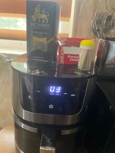 This image shows a close-up of a countertop setting, likely in a kitchen, with several objects visible. A black appliance, possibly an air fryer or a multi-cooker, dominates the right side of the frame, its digital display showing "03". The appliance has several buttons and labels, including "МЕНЮ", "МИН", "ВРЕМЯ", and an icon of a potato, possibly indicating a "fries" or "potato" function. There are some white powdery residues on and around the appliance, suggesting it may have been recently used or is due for cleaning.
To the left of the appliance, a box of "RICHARD ROYAL TEA - ROYAL CEYLON" is prominently displayed. The box features a gold lion emblem and text in English. Further to the left, a red box with Russian text, including "Русск" (Russk) and "саха" (sakha), is partially visible. This box contains what appear to be wooden skewers, capped with a yellow lid. In the upper left corner, a stack of cutlery, including forks and spoons, is held in a metal container.
The overall scene suggests a domestic setting. The lighting appears to be artificial, possibly from an overhead light, casting soft shadows. There are no people visible in the image. The background shows wooden elements, possibly a table or cabinets, and a window with a view of greenery, hinting at an indoor setting during daylight hours. The image is oriented vertically, as if taken with a phone. The visible text includes product names, menu options, and potentially ingredients or instructions in Russian and English.