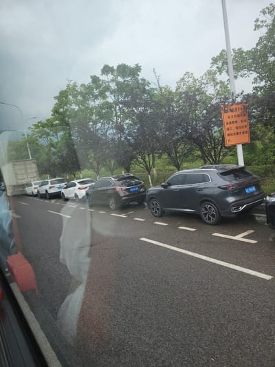 A queue of vehicles, including white sedans, white SUVs, a dark SUV, and a dark sedan, are stopped on a wet asphalt road. The vehicles are lined up on the right side of the road. In the background, there are trees and a cloudy sky. An orange sign with Chinese text is visible on the right side of the road, indicating road construction and a detour. The scene appears to be a highway or a main road, likely experiencing traffic congestion due to the road closure. The location is identified as Yufengshan, China.