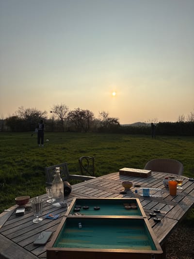 The image depicts an outdoor leisure scene in Bû, France, during late afternoon or early evening, characterized by the low sun casting a warm glow. In the foreground, a slatted wooden outdoor table is present, oriented diagonally. On the table, an open backgammon board with green felt and wooden frame displays black and light-colored checkers and two white dice. To the left of the board, a white mobile phone, a clear stemmed glass, a small patterned bowl, a cigarette packet, and a lighter are arranged. Behind the backgammon board, a clear glass bottle of water and another clear stemmed glass are visible. To the right of the board, a small plastic water bottle, a bowl containing items, an orange plastic cup, and a clear stemmed glass with an amber beverage are placed. A woven wicker chair and a wooden chair are partially visible around the table.
The midground features a expansive green grass lawn. Two individuals are engaged in activity on the lawn. One person, positioned to the left of center, wears a dark jersey with the number "10" on the back and dark shorts/pants, captured in the act of kicking a white and black soccer ball, which is airborne to their left. A second person, dressed in dark clothing, stands further to the right near a dense green hedge line that forms the boundary of the property. Several deciduous trees with bare branches are scattered across the midground.
The background shows a clear sky transitioning from pale blue overhead to a warm orange-yellow near the horizon. The sun, a bright yellow-orange orb, is prominent in the upper right quadrant of the sky. A faint line of distant, low-lying hills or landscape features is visible beneath the sun.
