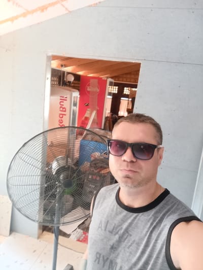 The image depicts a man wearing sunglasses and a grey sleeveless shirt. He is standing in what appears to be a construction or renovation site, with unfinished walls and exposed wooden beams visible. A large black fan is positioned to his left. In the background, through an open doorway, a red Coca-Cola branded refrigerator and a white refrigerator are visible. The scene is set in a brightly lit indoor environment. While the image contains visual cues related to a city, it does not provide any specific landmarks or details that would allow for a precise location within Ladyzhyn, Ukraine.