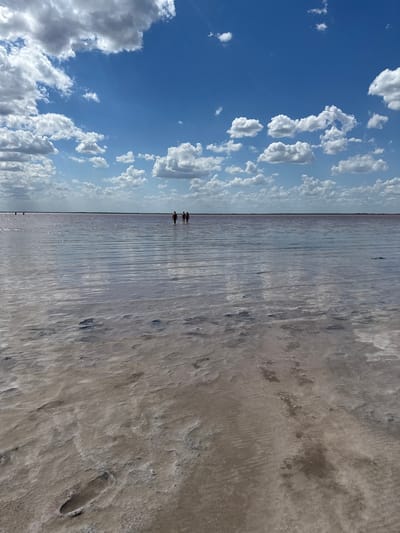 This is a wide shot of a shallow, pink-tinged salt lake under a bright blue sky dotted with fluffy white clouds. The water is calm and reflects the sky, creating shimmering horizontal lines. The foreground shows a sandy, slightly textured bottom with visible footprints. In the middle distance, three people are wading in the water, appearing as dark silhouettes. The horizon is a thin line separating the water from the distant, flat land. The overall impression is one of open space and a serene natural environment. The location is identified as the salt lake in Bursol', Russia.